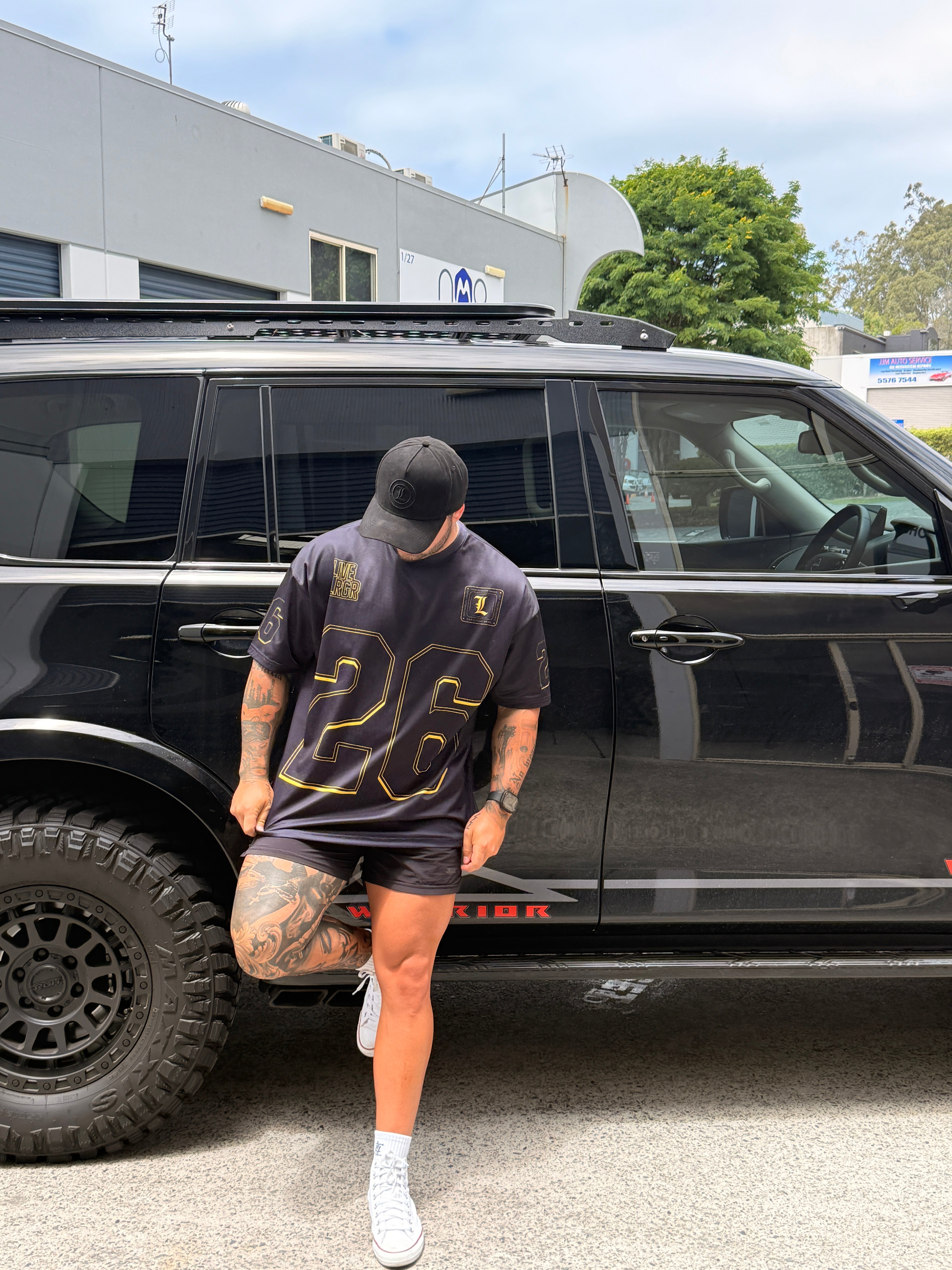 Person wearing a LRGR black t-shirt with gold text, standing next to a black vehicle.
