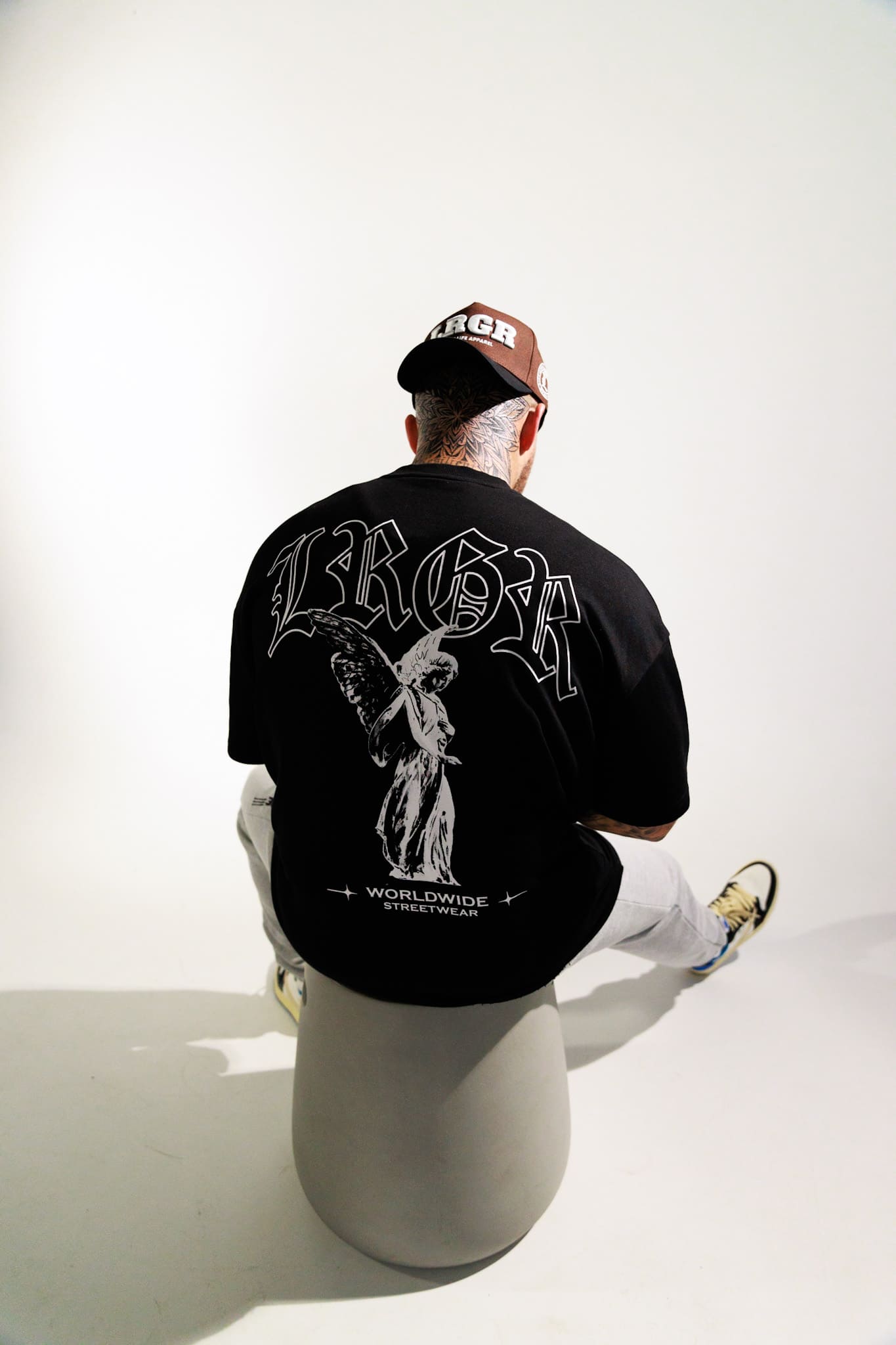 Person wearing a black t-shirt with white graphics and text, sitting on a white stool against a white background.