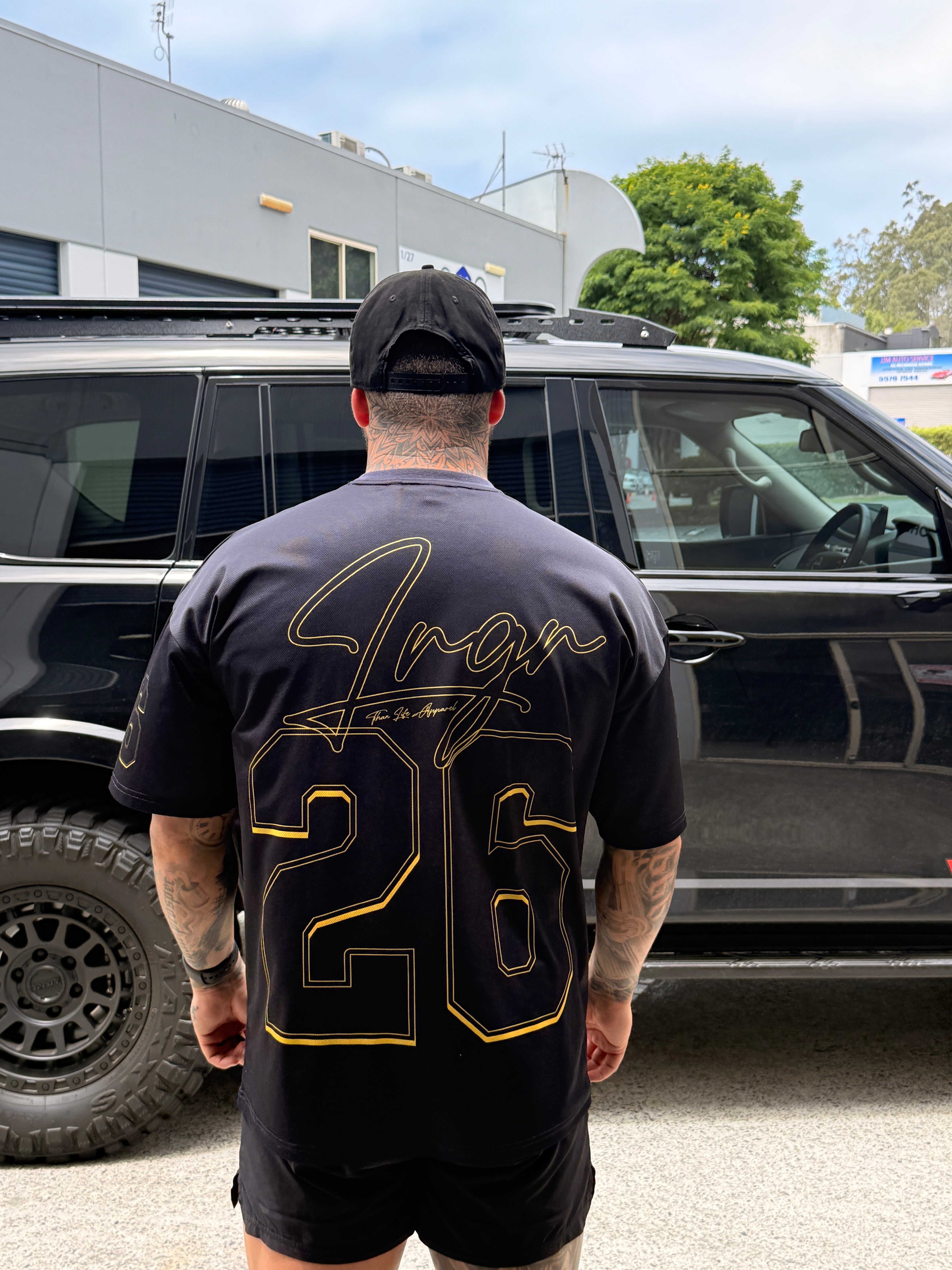 Person wearing a LRGR black t-shirt with gold text, standing next to a black vehicle.
