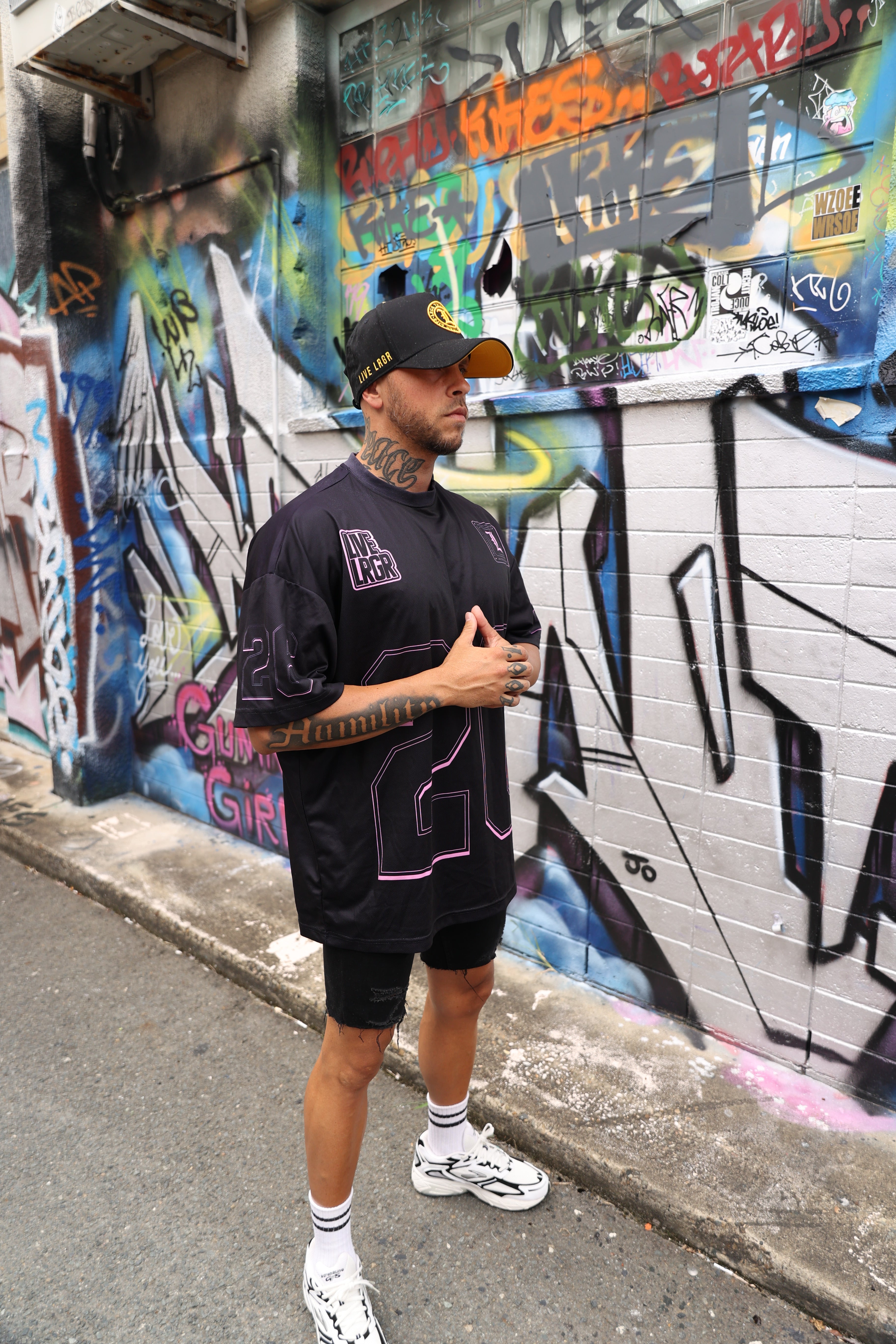 Person leaning against a colorful graffiti wall on a street, wearing a black-pink LRGR t-shirt