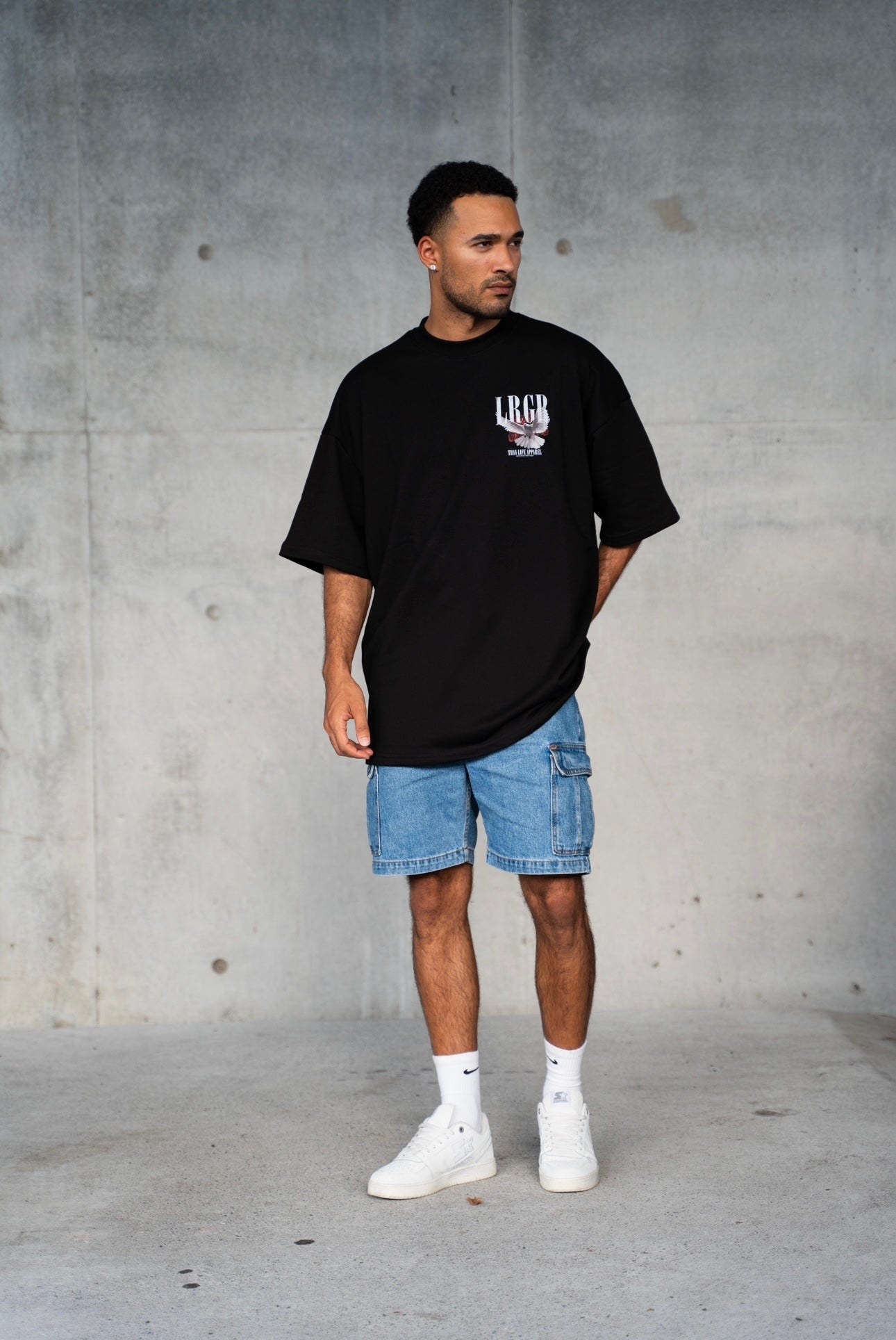 Person wearing a black t-shirt with a logo and text, standing against a concrete wall.