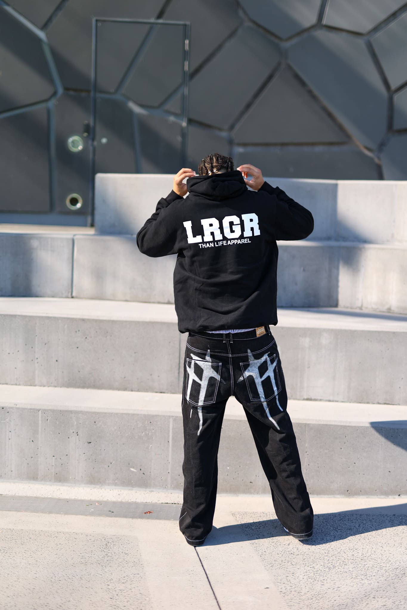 Person wearing a black hoodie with 'LRGR' branding on steps against a geometric wall.