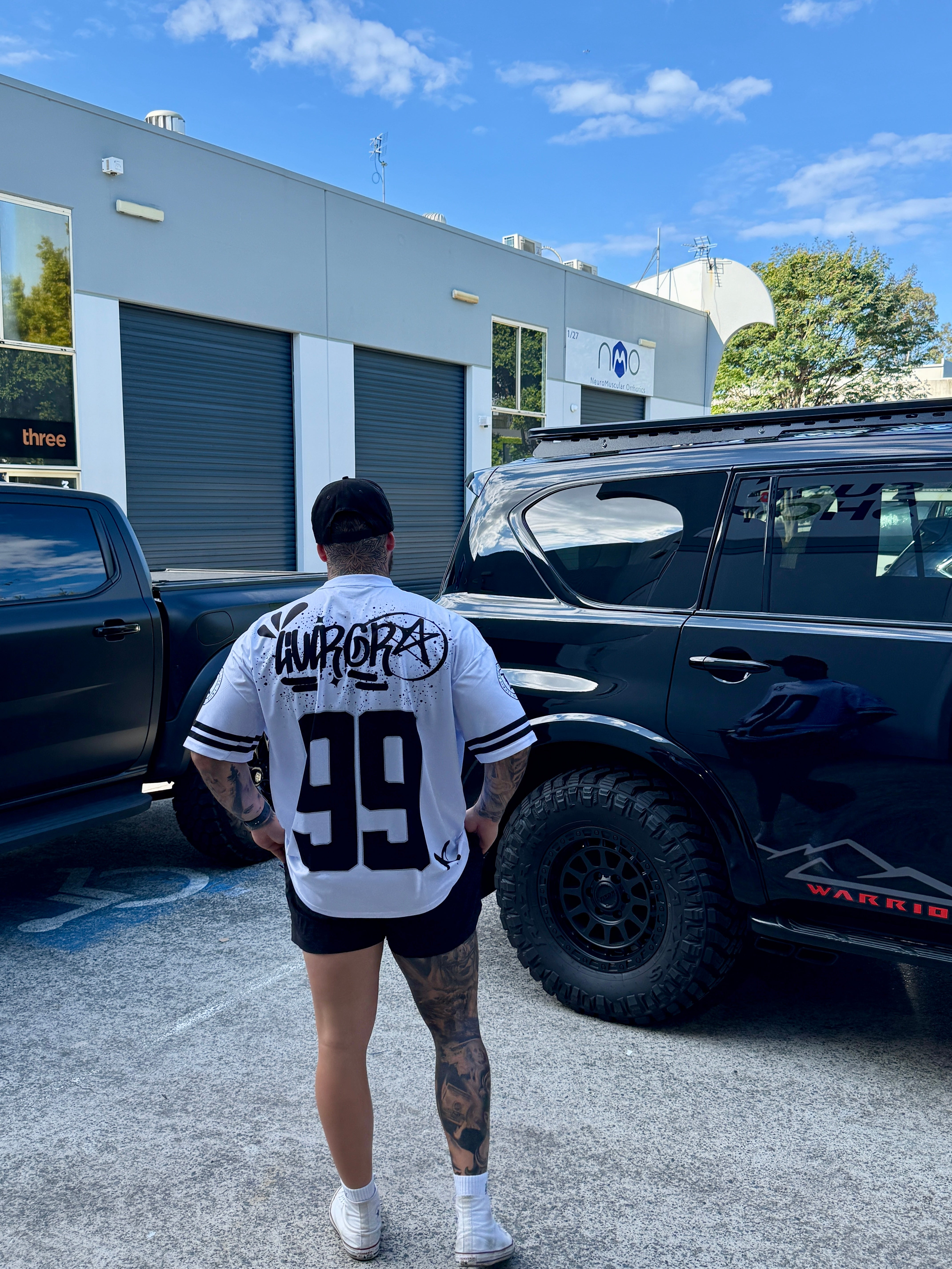 Person wearing a white t-shirt with black text and numbers, standing in front of two black vehicles.