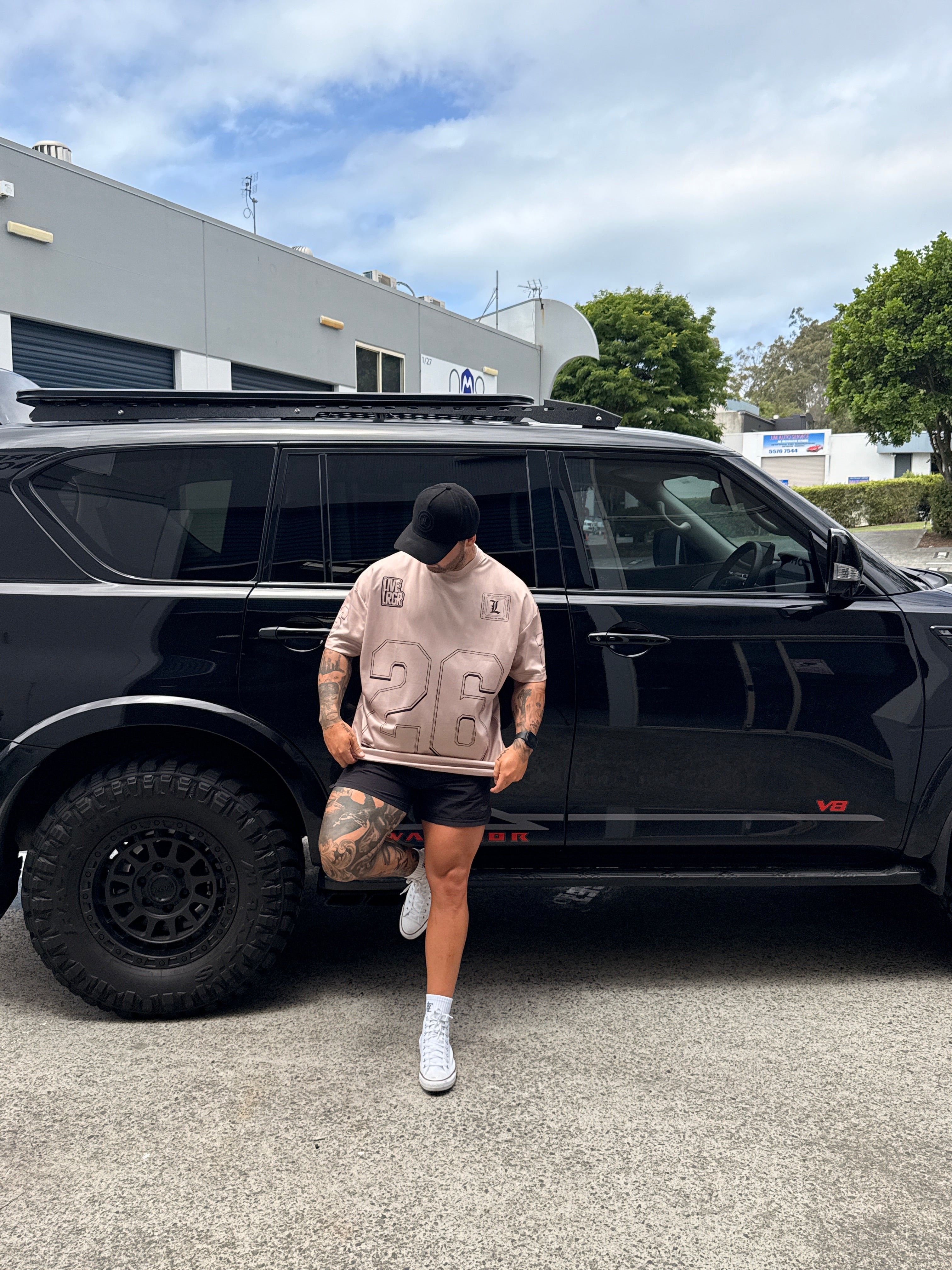 Person wearing a LRGR beige t-shirt with a logo, standing next to a black SUV on a street.