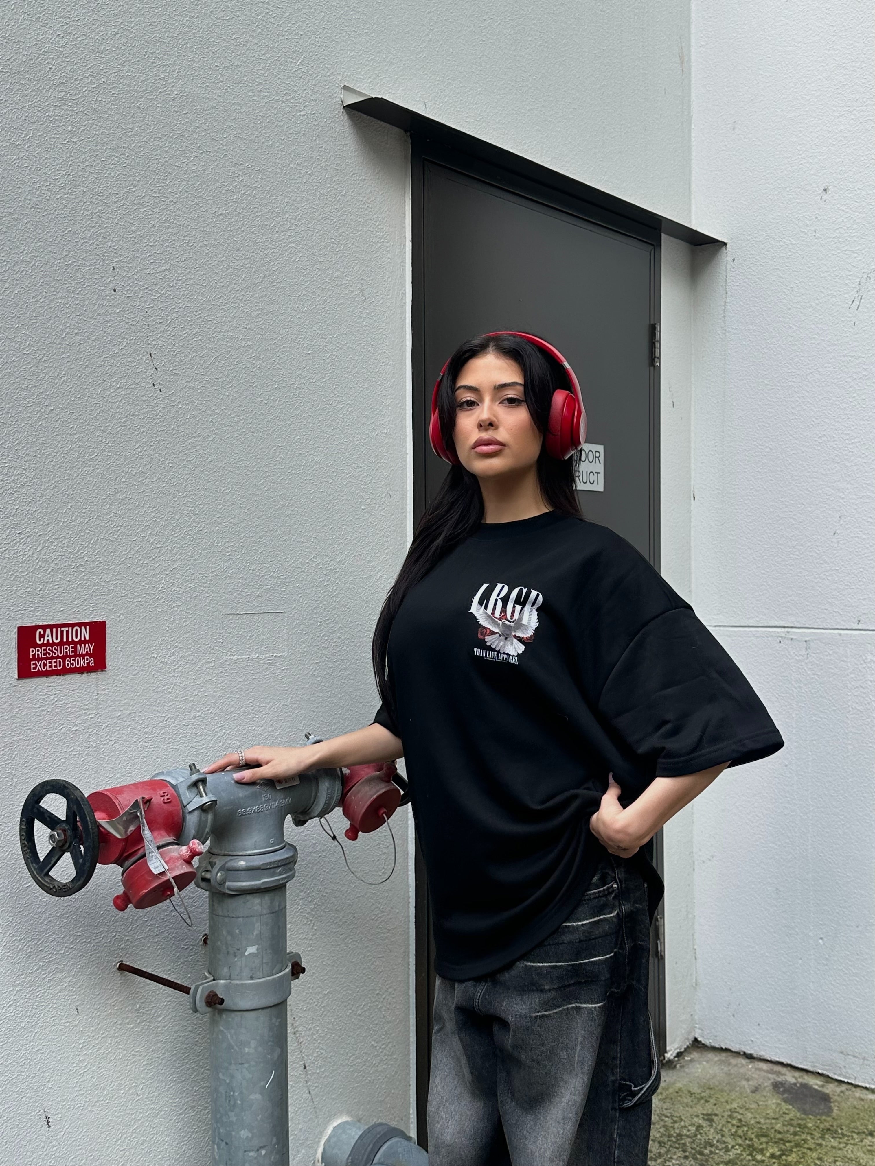 Person wearing a black t-shirt with a logo and text, standing against a concrete wall.