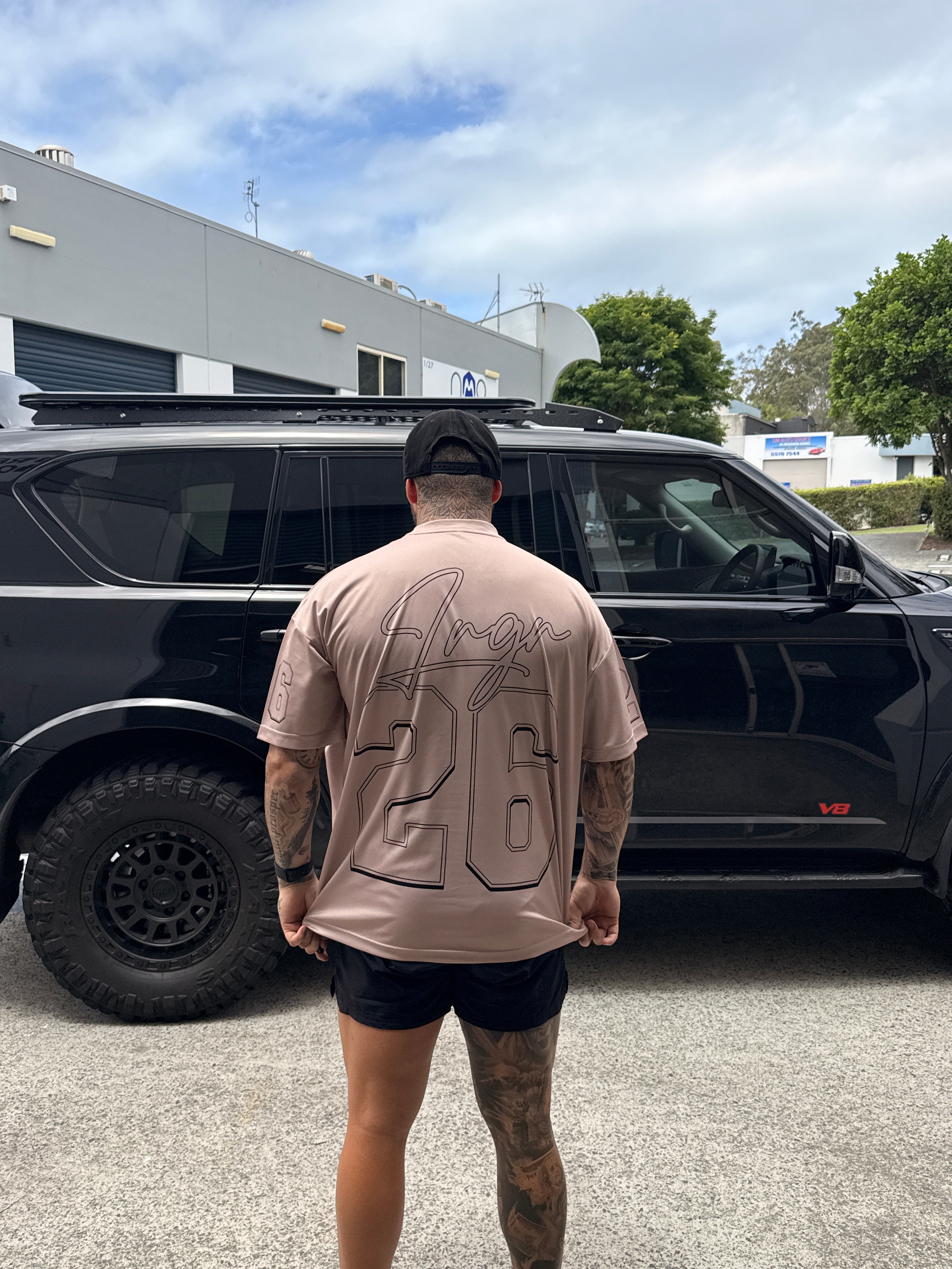Person wearing a LRGR beige t-shirt with a logo, standing next to a black SUV on a street.
