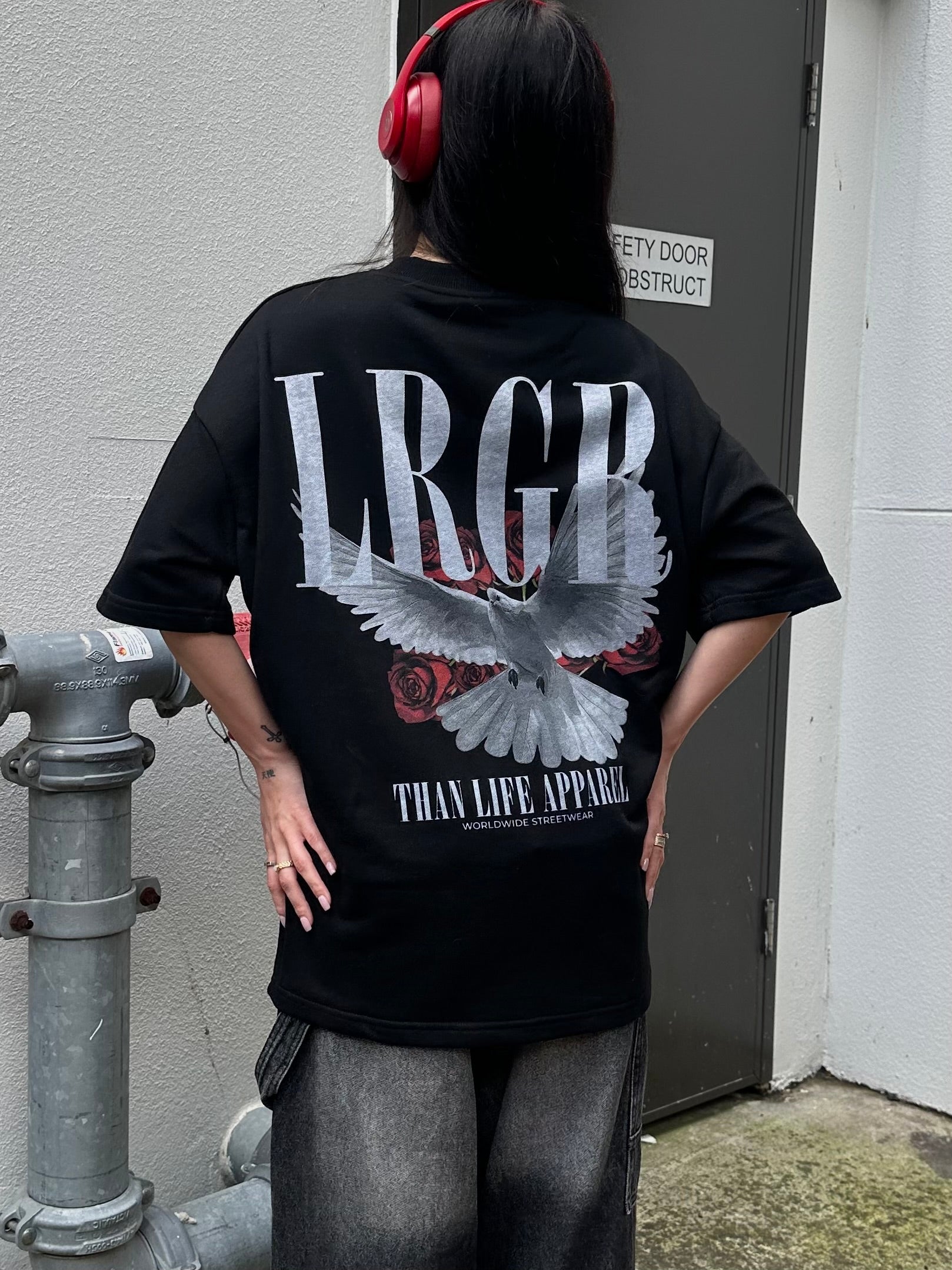 Person wearing a black t-shirt with a logo and text, standing against a concrete wall.