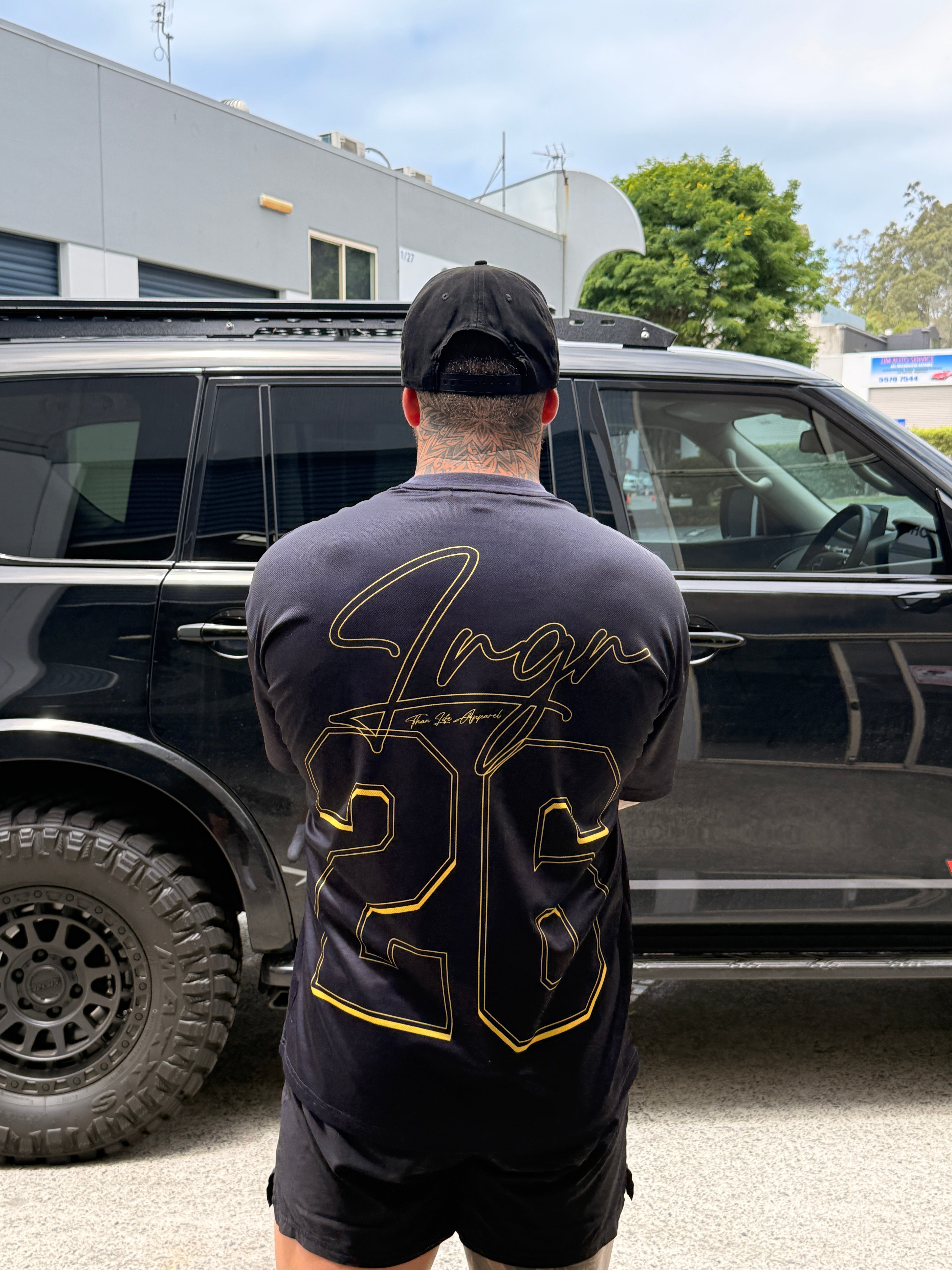 Person wearing a LRGR black t-shirt with gold text, standing next to a black vehicle.