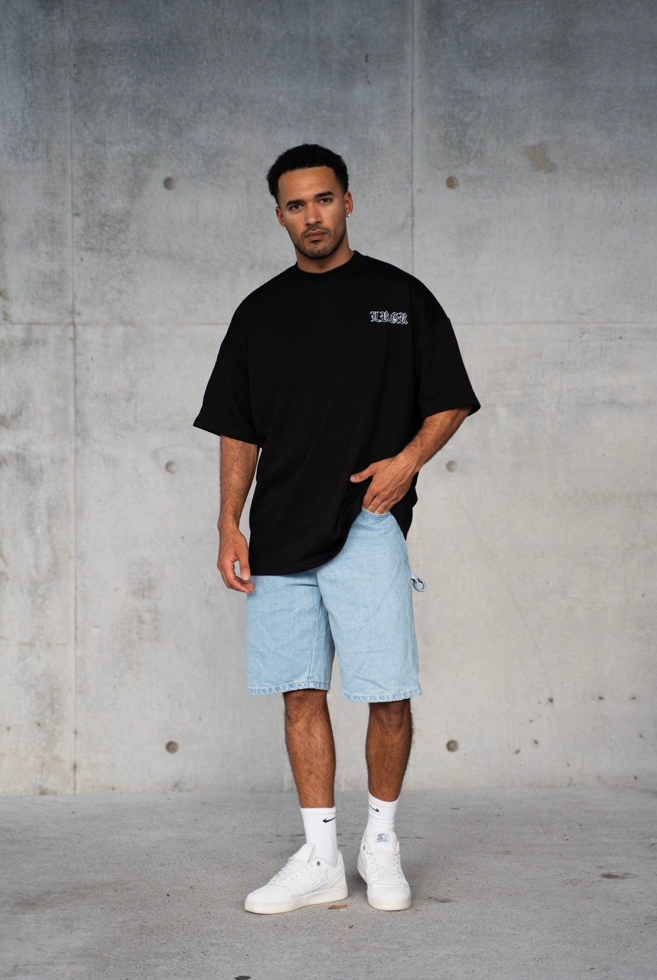 Person wearing a black t-shirt with white graphics and text, sitting on a white stool against a white background.