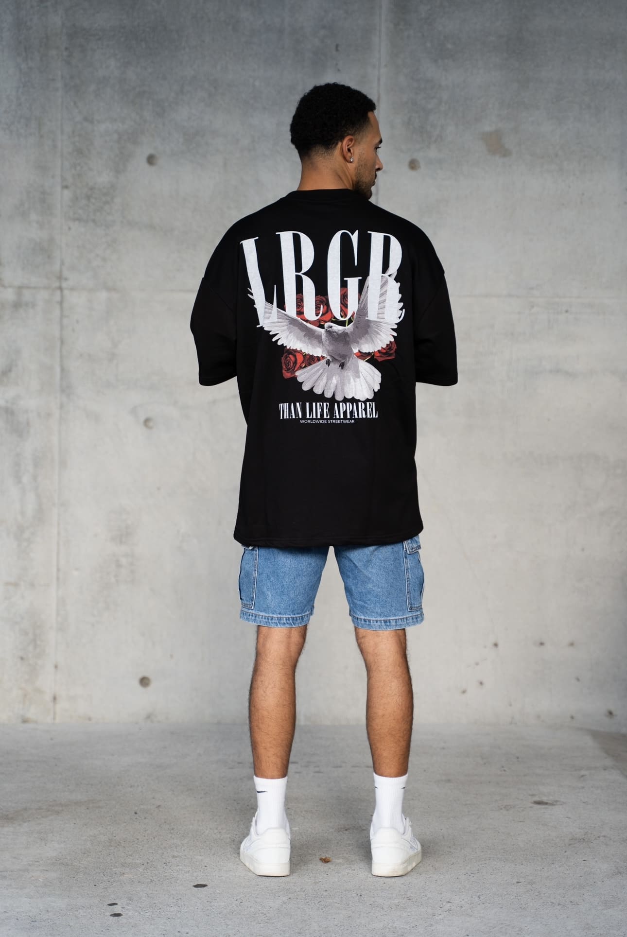 Person wearing a black t-shirt with a logo and text, standing against a concrete wall.