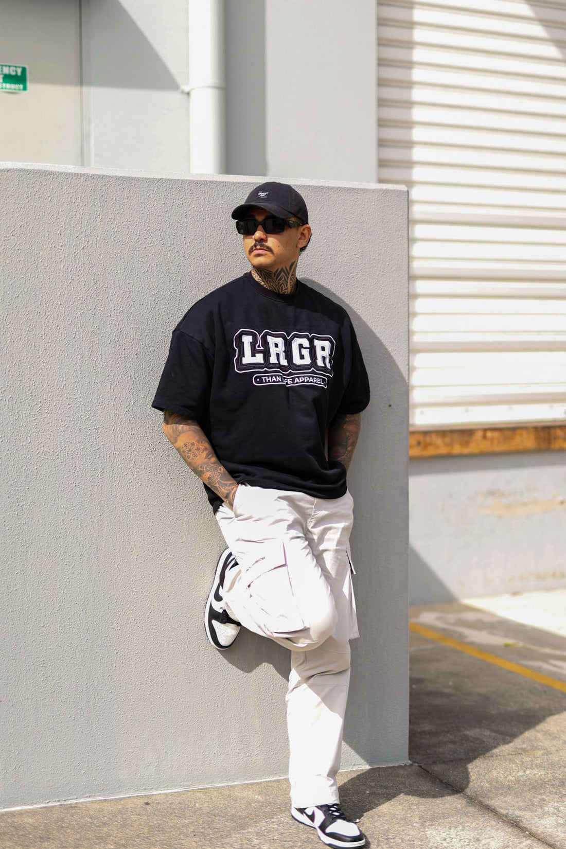 Person wearing a black 'LRGR' t-shirt and white pants leaning against a wall.