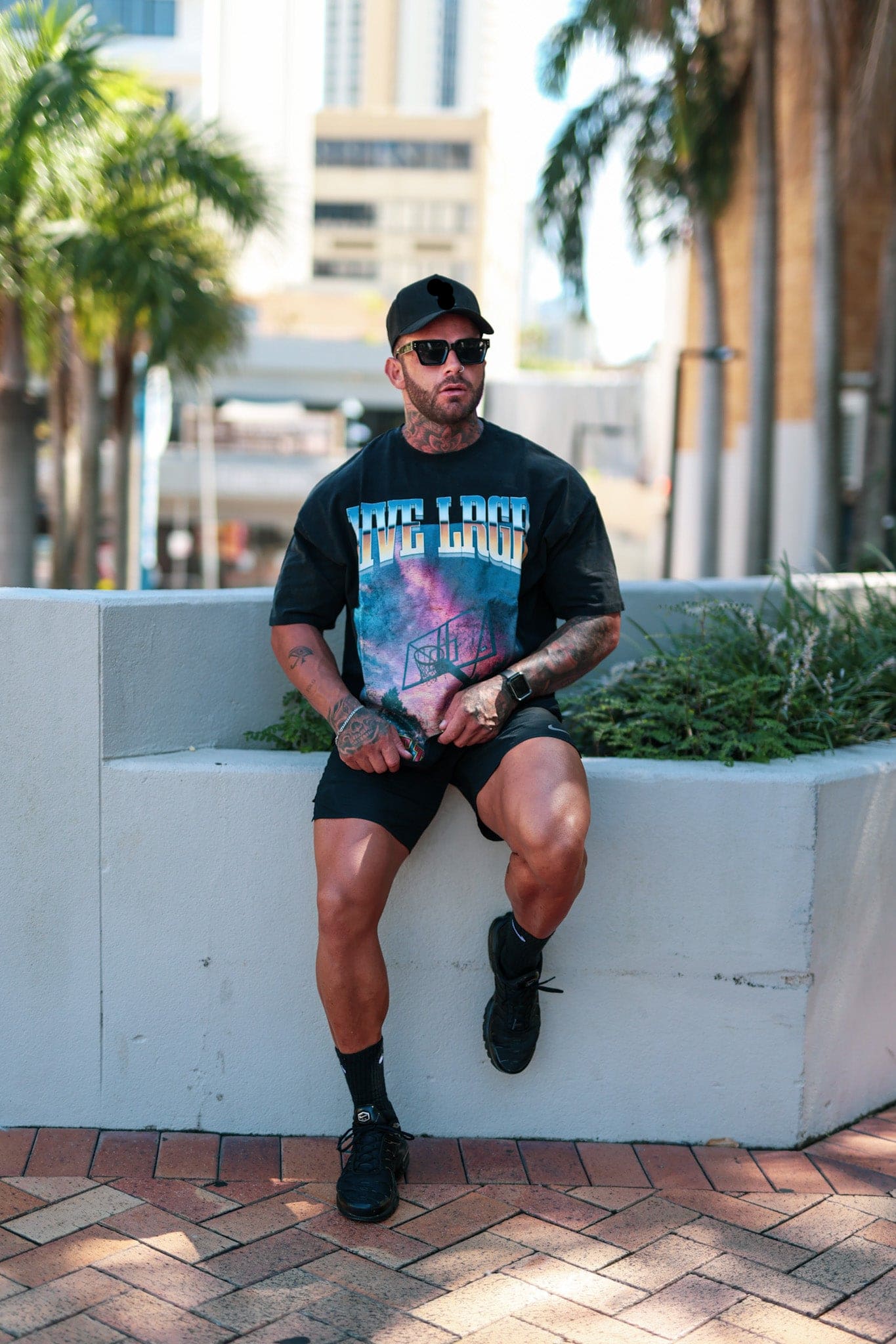 Man sitting on a ledge wearing a LRGR graphic t-shirt and sunglasses, with a cityscape background.