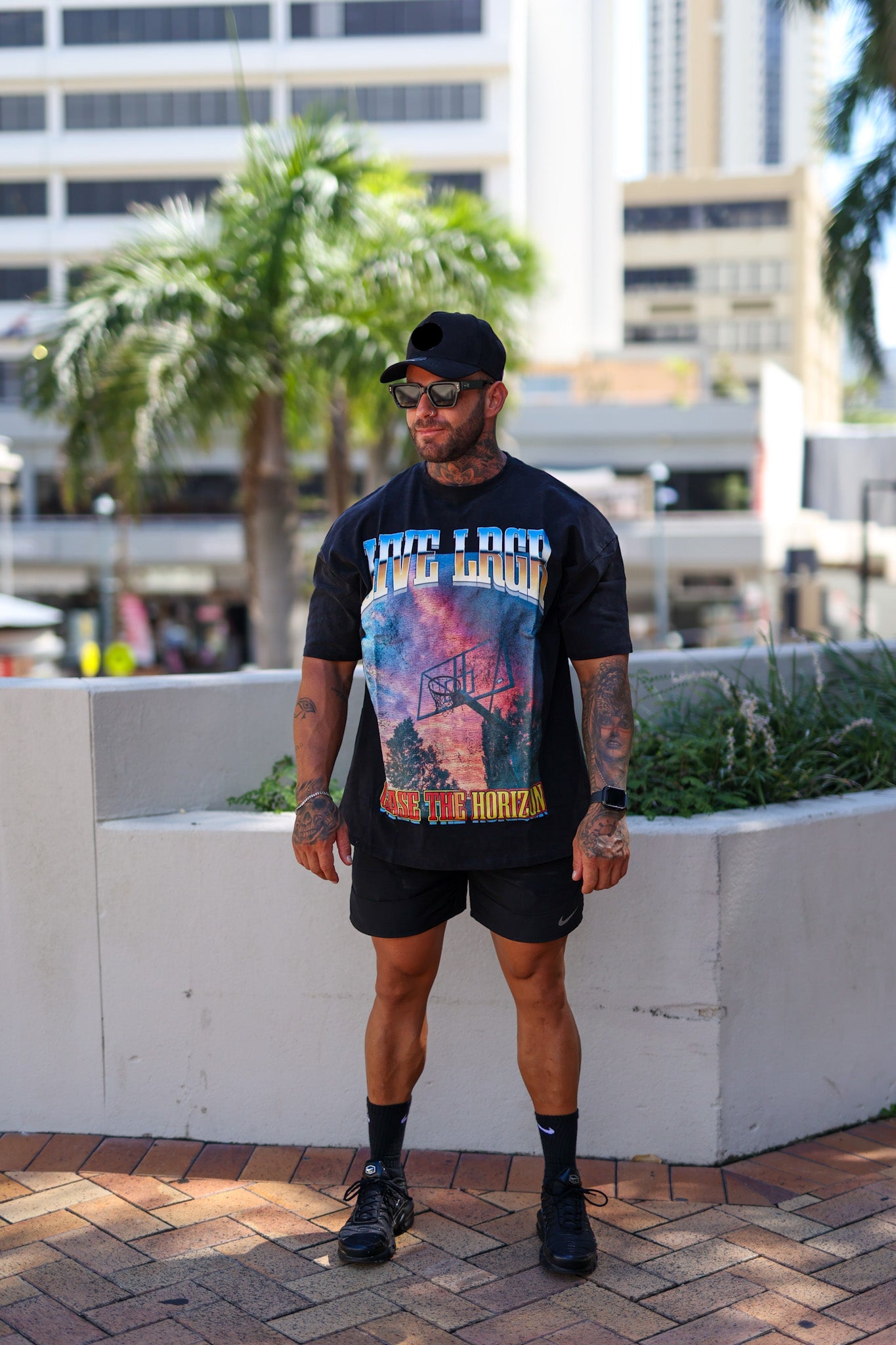 Man sitting on a ledge wearing a LRGR graphic t-shirt and sunglasses, with a cityscape background.