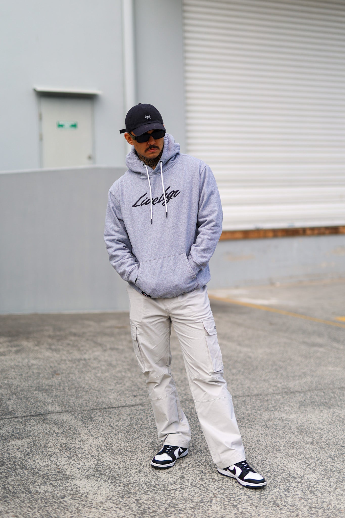 Man wearing a gray hoodie and sunglasses, leaning against a wall.