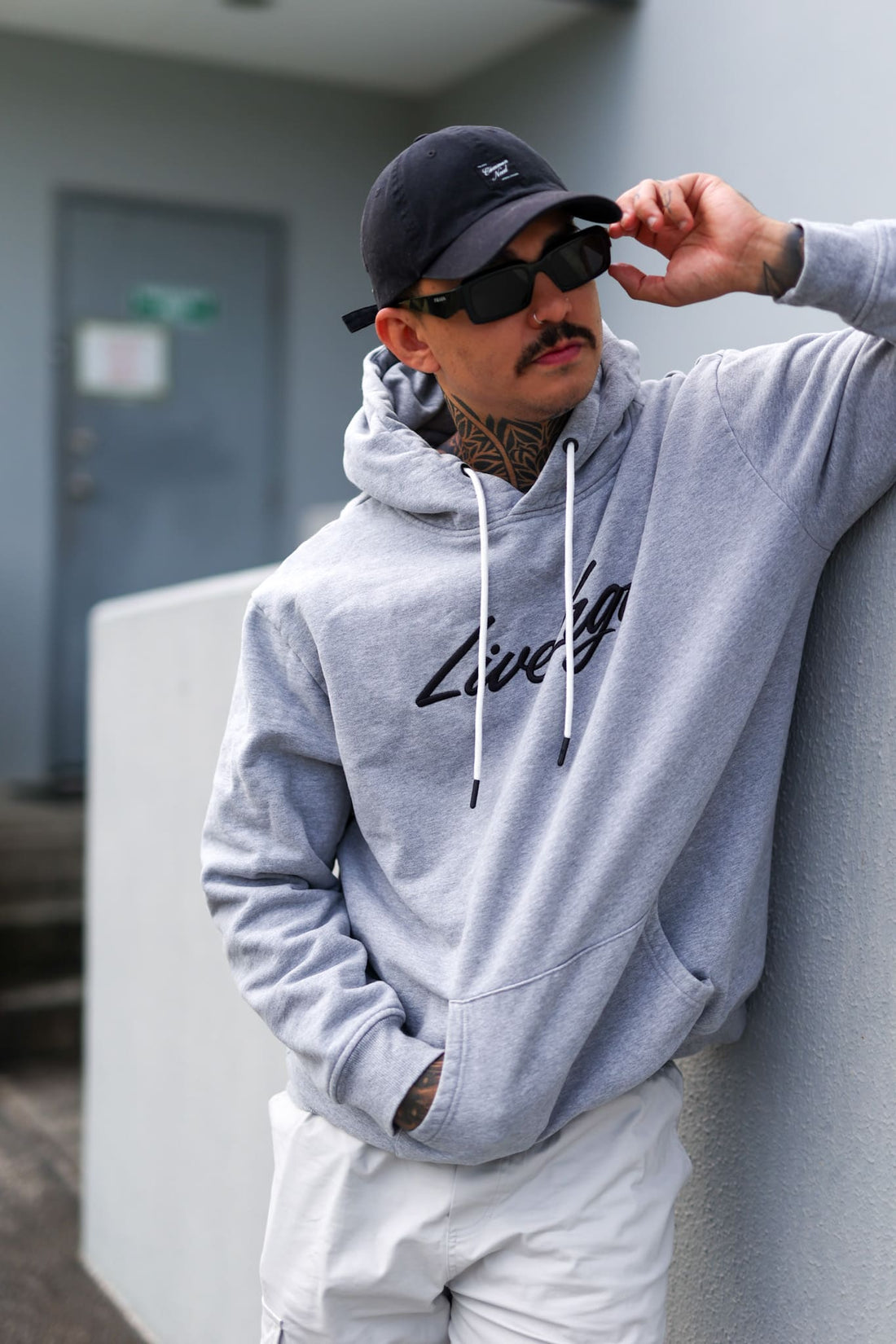 Man wearing a gray hoodie and sunglasses, leaning against a wall.