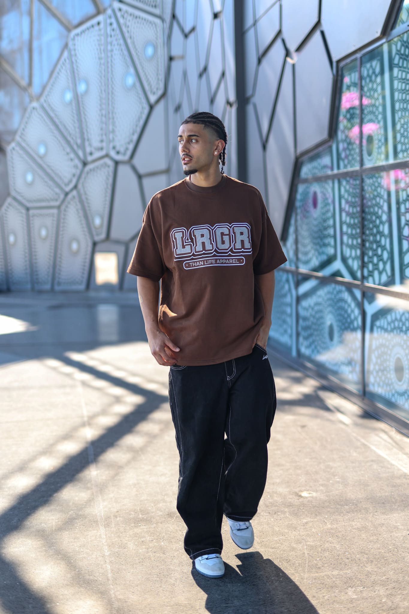 Person wearing a brown t-shirt with 'Laga' printed on it, standing in front of a modern architectural structure.